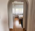 A narrow hallway with wooden flooring and light-colored walls, leading to a bedroom with a bed, rug, and window with curtains.