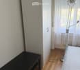 A small, furnished room with a wooden floor, a black table, a white wardrobe, and a floor lamp near a window with sheer curtains.