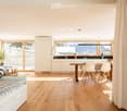 Open-plan living and dining area with light wood flooring, white walls, and large windows overlooking a snowy mountain view. Features a sofa, dining table with chairs, kitchen area with cabinets and a sink, and a potted plant near the window.