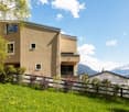 Modern two-story house with beige facade, large windows, and a raised ground floor, surrounded by a green lawn and trees with mountain views in the background.