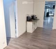 Bright hallway with wooden flooring, white walls, and a small coffee station with a table and shelves. An open doorway leads to another room with visible furniture.