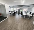 Modern office space with wooden flooring, large windows, white tables, black chairs, and privacy screens.