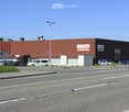 A large industrial-style building with a red metal roof and signage for "FIRSTCARS" visible. The building has multiple entrances and a parking area with several cars.