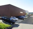 A large industrial-style building with a brown metal facade and a flat roof, surrounded by a parking lot with several cars.