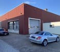 A large industrial-style building with a red metal facade, large garage door, and several windows. Several cars are parked in front of the building on a paved area.