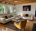 Bright living room with wooden floors, large windows, two sofas, coffee table, armchair, floor lamp, and a piano in the corner.