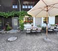 Outdoor patio area with stone pavement, large umbrella, dining table, and chairs, surrounded by greenery and a house with wooden and white walls.