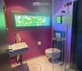 Modern bathroom with pink walls, white tiled floor, a sink, toilet, and a glass-enclosed shower. A large window provides natural light, and a towel hangs on the wall.