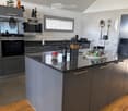 Modern kitchen with grey cabinets, black granite countertop, stainless steel appliances, and various kitchen items including a knife block, wine bottle, and decorative items on the counter.