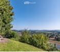 A serene garden or outdoor area with a table and chairs, surrounded by lush greenery and offering a panoramic view of a town and distant hills.