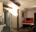 Cozy bedroom with a bed, red bedding, a chair, a table, a mirror, a floor lamp, and a television. The room features exposed wooden beams and a small seating area.