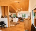Open-plan living and dining area with wooden floors, exposed wooden beams, large windows offering a view, a white sofa, coffee table, kitchen island, dining table with chairs, chandelier, speaker, and decorative items.