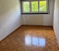Empty room with wooden parquet flooring, white walls, and a large window with three panes showing greenery outside. A radiator is mounted on the wall below the window.