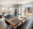 Modern kitchen with a light grey color scheme, featuring a central wooden dining table surrounded by blue chairs, a kitchen island with a countertop, and built-in cabinets with stainless steel appliances.