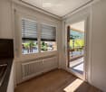 Bright room with wooden flooring, radiator, large windows with blinds, and a sliding door opening to a balcony with outdoor view.