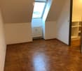 Empty attic room with wooden parquet flooring, white walls, and a skylight window. A radiator is visible near the window, and built-in shelving is on the right side.