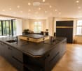 Modern kitchen with dark countertops, stainless steel sink, island, and built-in cabinets. Large windows and a dining area with chairs and a table are visible.
