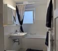 Modern bathroom with a white sink, bathtub, toilet, mirror, radiator, and a window with curtains. Towels are placed on the radiator and floor.