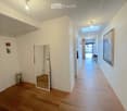 Bright hallway with wooden flooring, white walls, and modern lighting fixtures. Features a mirror, small decorative table, and artwork on the walls.