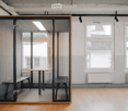 A modern, minimalist room with wooden flooring, large windows, and a glass partition featuring a small table and bench inside.
