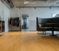 Modern, minimalist room with light wooden flooring, a large L-shaped sofa, a small round table, a wooden shelf, and a coat rack with jackets.