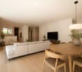 Modern living and dining area with light-colored furniture, a large sectional sofa, a round wooden dining table with chairs, a television, and decorative plants.