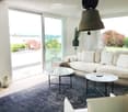 Bright living room with large windows and a balcony view, featuring a cream-colored sectional sofa, coffee table, side table, and a patterned rug.