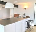 Modern kitchen with white cabinets, a central island, and a built-in stove. Features include a hanging pendant light, a wall-mounted fan, and two stools at the island.