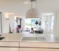 Modern kitchen with white cabinets, a large island, and a view of the ocean through large windows. The space includes a dining area with a table and chairs, a fan, and decorative vases with flowers.
