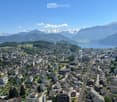 Aerial view of a town nestled in a valley with surrounding mountains and a lake in the background. The town features a mix of residential and commercial buildings, roads, and green spaces.