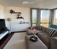 Modern living room with a grey sectional sofa, coffee table with a flower vase, floor lamp, TV stand, and decorative shelves with plants and a clock.