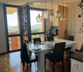Modern dining area with a white dining table, six black chairs, pendant lights, and a laptop on a nearby desk. Large windows offer a scenic view.