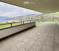 Spacious balcony with modern tiled flooring, expansive views of a lake and surrounding mountains, glass railing, and a grassy area.