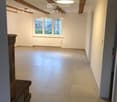 Empty room with exposed wooden beams, tiled floor, large window, and modern lighting fixtures.