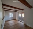 Empty room with wooden floor, exposed wooden beams on the ceiling, white walls, and a large window with white frames. A sliding glass door leads to a balcony or terrace area.