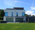 Modern two-story house with light blue exterior, large windows, covered patio area, outdoor seating, grill, and hot tub in a well-maintained lawn.
