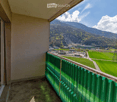 Balcony with green railing, overlooking a scenic valley with mountains, fields, and scattered buildings.