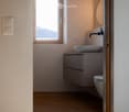 Bathroom with wooden floor, white walls, a window, a sink, a toilet, and a mirror
