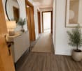 Modern hallway with wooden flooring, white walls, and a slanted wooden ceiling. Features include a round mirror, a small table with a lamp, a potted plant, and a framed artwork on the wall.