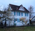 Two-story white house with blue shutters, red-tiled roof, and small garden area with trees and a fence.