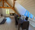 Loft-style room with wooden beams, fireplace, sofa, dining table with chairs, and a fan. Large windows provide natural light.
