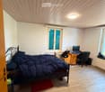 Cozy attic bedroom with wooden floors, slanted wooden ceiling, large windows with shutters, a bed with dark bedding, a desk with a computer, and a chair.