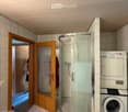 Bathroom with a glass-enclosed shower, a washing machine, and a wooden door leading to another room.