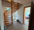 Spacious hallway with wooden flooring, a wooden spiral staircase leading upwards, and a small metal table with items on it. A doorway leads to another room with visible furniture and items.