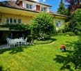 Lush garden with a well-maintained lawn, a patio area with a white table and chairs, and a large house with yellow and red exterior walls and multiple windows.