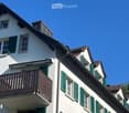 Two-story white building with green shutters, multiple windows, and a wooden balcony on the second floor.