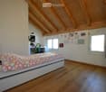 Bright bedroom with wooden floors and slanted wooden ceiling. Features a single bed with colorful heart-patterned bedding, a small desk with a chair, and a mirror on the wall.