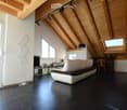 Modern living room with exposed wooden beams, large windows, and skylights. Features a white sectional sofa, a wooden chair, a small table, and a television on a stand.