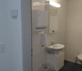 Modern bathroom with a white sink, faucet, toilet, paper towel dispenser, mirror, and a small trash bin.