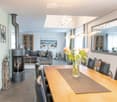 Open-plan living and dining area with a grey sectional sofa, wooden dining table with black chairs, modern fireplace, large mirror, and decorative vases with flowers.